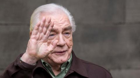 Brian Cox looking over to the right waving to someone. He is smiling and wearing a green shirt and brown jacket.