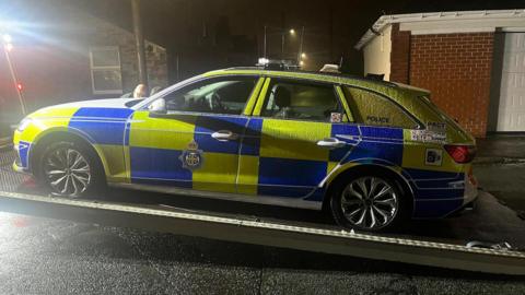 A police car being placed on to a tow truck ramp on a road. It is night time and it has been raining.