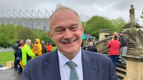 Sir Ed Davey is smiling and wearing a blue suit, patterned tie and pale shirt. There are people behind him and Newcastle United's football stadium.