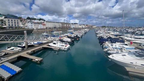 Rows of boards moored next to pontoons in a harbour on a cloudy day, with a series of buildings running behind them.