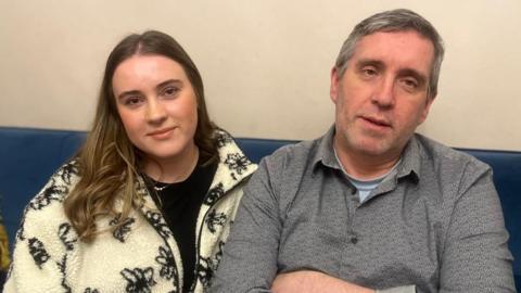 Aimee sitting on a sofa with her dad Damien. Aimee has long brown hair with blonde ends, she's wearing a black top and a cream zip up cardigan with black flowers. Damien has light grey short hair and is wearing a grey shirt with a light blue top underneath.