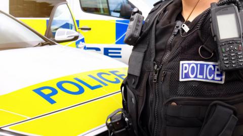 The images shows the front of a police car and an office standing next to it, with a strip saying "police" on the right-hand-side behind a hand-held radio. There is also another police car behind the police car.
