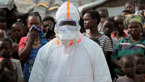 A health worker in personal protective equipment (PPE) stands near displaced people waiting for the burial of suspected Ebola victims at the Kigonze displaced persons camp, one month after an outbreak was declared, in Bunia, eastern Democratic Republic of Congo