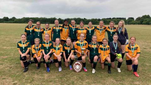 A team of young girls pictured with their trophies on a football pitch. The girls are arrange in two rows with one row standing and the other kneeling. The kits are yellow and green.