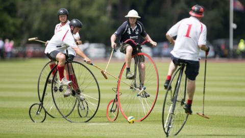 Four players ride penny farthing bicycles across a grass pitch, striking a yellow ball with wooden mallets