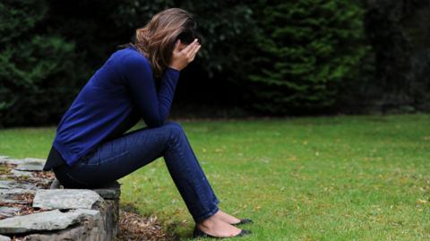 A woman dressed in blue with her head in her hands, while sat outside in a garden