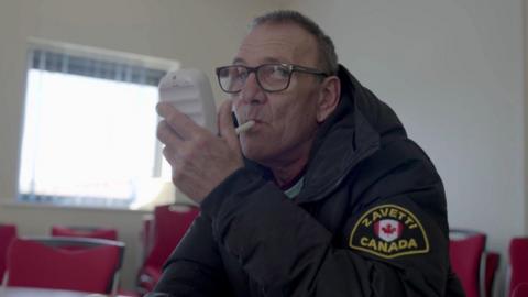 A screenshot from the 'Quitters' which shows Andy, a man who looks to be in his 60s, wearing glasses with a black frame. He is holding a small white machine in his hand, with a tube in his mouth as he blows in to it. He is white, has short grey hair and is wearing a black puffer coat with a Canada badge on the left sleeve. He is sitting in a room with red chairs and a large square window in the background. 