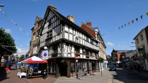 The corner of a two-storey black and white timbered building with pedestrianised streets running down both sides and market stalls with red and white awnings on the left hand side. There is bunting hanging over both streets.
