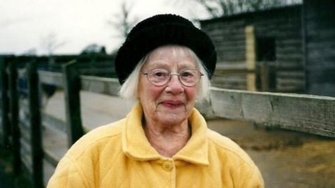 An elderly woman wearing a yellow button up coat and a black hat.