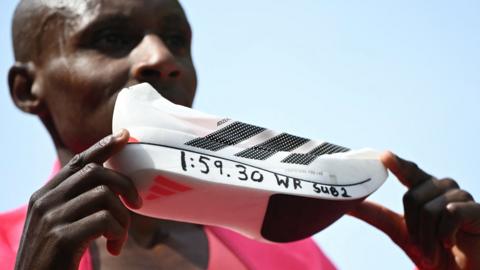 A white Adidas running shoe is held up so that its side is central to the image. It has the three black stripes on it for the Adidas logo and the text 1:59:30 WR sub 2 is written along the base of the sole. The two hands of marathon world record holder Sabastian Sawe can be seen grasping the shoe lightly between fingers, with Sawe's blurred face, covered by the shoe, is partially seen in the background