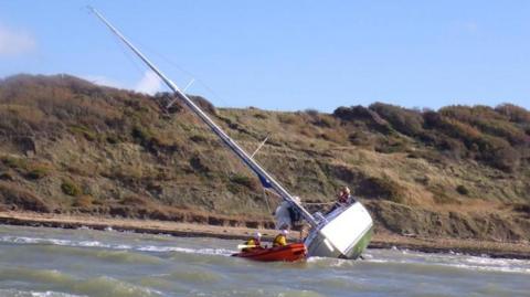 A yacht tilts alarmingly in the sea near the Isle of Wight coast. An RNLI dinghy with two crew waits in the water nearby.
