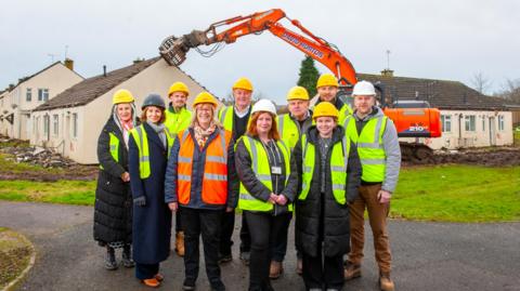 A group of people wearing hard hats, high vis vests and winter coats stand in a group and smile at the camera. Heavy machinery can be seen demolishing a bungalow, amid other houses, in the background. 