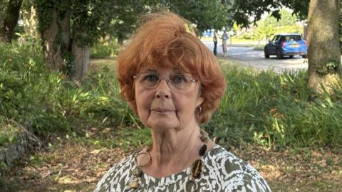 A woman with red hair and glasses wearing a green and white pattern top standing in a grassy area with trees in the background 