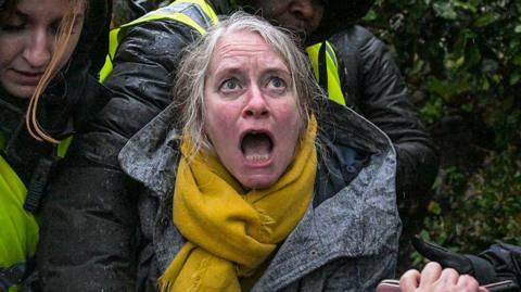 A close up of a woman with grey hair with her mouth open in horror. She is wearing a mustard yellow scarf.