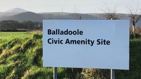 A large white sign that reads Balladoole Civic Amenity Site in black lettering in front of a field, with hills in the background on a clear day.