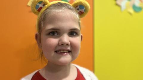 A little girl smiles straight at the camera. She has blonde hair tied back and is wearing a yellow fluffy headband with teddy-bear style "Pudsey" ears. She has dark brown eyes. She is wearing a red top and white cardigan. The background is bright and colourful with blocks of orange and yellow.