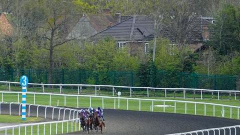 Horses and riders rounding a bend on a racetrack at Kempton Park, with green fencing, trees and houses visible beyond the course