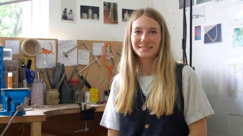 The image shows Iona Hall wearing a white t-shirt underneath a denim pinafore, with silver chains layered around her neck. She has long blonde hair and is smiling at the camera, sitting at a desk full of tools used to make jewellery.