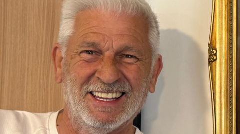 Stewart Woods, with white hair and a short white beard, smiles for the camera. He's wearing a white T shirt and is standing in front of a wall with the edge of a gold picture or mirror frame visible.