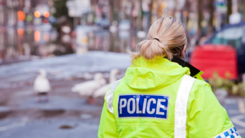 A female officer in hi-vis yellow police emblazoned uniform walks down a road in a city or town centre. She has her back to the camera but a flock of swan or geese can be seen in the distance with the remaining view blurred out.