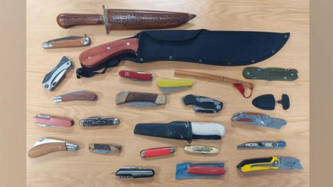 Twenty-four knives of various sizes resting on a table. Some are large, some are small and many are pocket knives. 