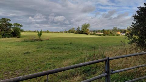 A green field behind a metal fence. There are some clouds in the sky but the sun is shining through.