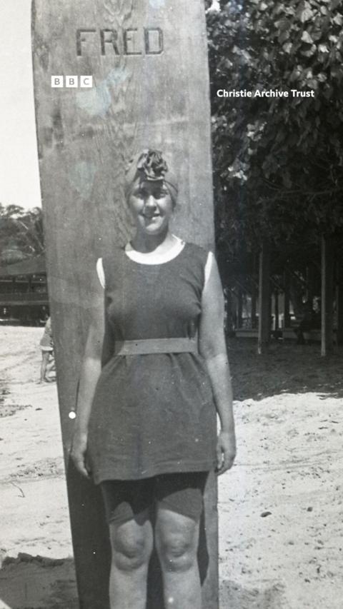 A young Agatha Christie stands on a beach against a large surf board.