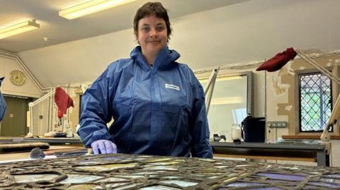 A woman with short dark hair wearing a blue protective jacket and blue gloves, stands beside a table covered with a piece of stained glass. Behind her there are windows and lamps with red cloths over them.
