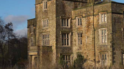 A 16th century manor house is surrounded by overgrown trees and shrubbery. A lot of its windows are broken. 