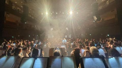 The view of an orchestra from the perspective of a musician. There is a row of music stands with sheet music on them in the foregournd, and an orchestra facing out towards an audience further away from the camera. There are bright lights near the top of the image shining onto the group of players.