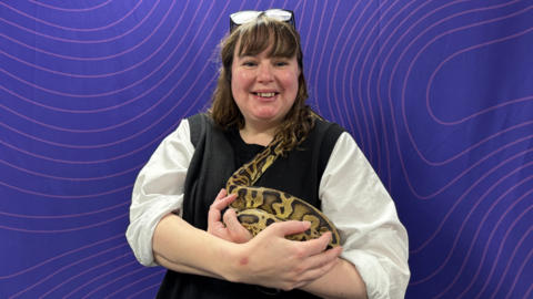 A woman, Vikki Buxton-Helyer, who is wearing glasses on top of her head and a white and black top, is holding a large yellow and black spotted snake in her arms. She is smiling and standing in front of a purple background.