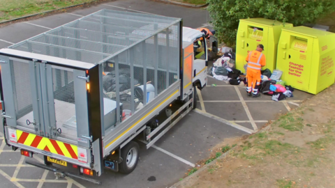 A council worker dressed in fluorescent suit standing over fly-tipped rubbish next to two charity bins.