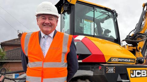 Mark Deaville standing front of a JCB tractor. He is wearing an orange and grey hi- vis jacket and a white hard hat. Under this he is wearing a jumper, white shirt and stripy tie. 