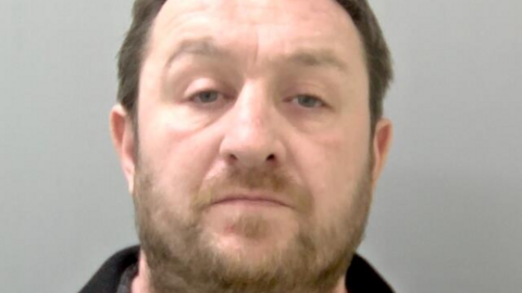 Scrase appears in a police mugshot in a dark-coloured top. He has brown hair and beard.