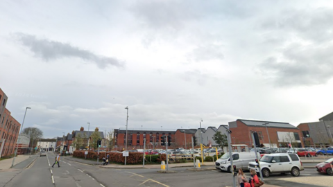 A man cross the road while the traffic light is green and two women are waiting to cross the road while holding bags. A silver van and White range rover are waiting at the traffic lights. There are red brick buildings with grey tiles in the background 