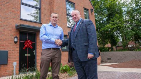 Two men shake hands outside a newly-built house which has a red ribbon tied to a black front door. There are trees and further houses in the background.