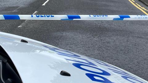 The white bonnet of a police van parked next to a blue and white police cordon stretched across a grey road.