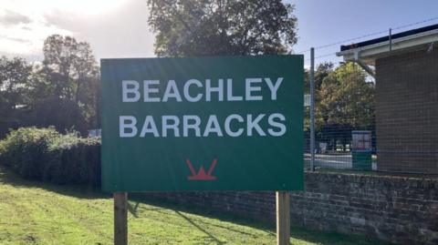 There's large green sign with bold white text that reads “BEACHLEY BARRACKS”. Below the text, there is a red emblem which represents military insignia. The sign is on a neatly mown grassy area with trees with green foliage, a brick wall, which is the entrance to the barracks.