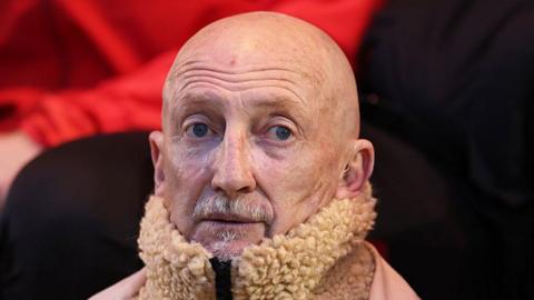 Swindon Town boss Ian Holloway looking towards the camera while sat down in the technical area.