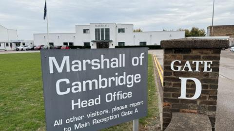 The entrance to Marshalls head office. A white building with three large windows at the front. There is a small car park to the front of the building and a sign that reads "Marshall of Cambridge Head Office". 