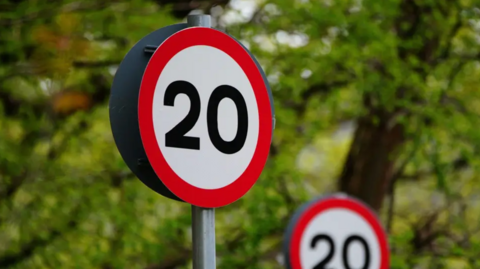 A close-up photograph of a circular red-and-white road sign that has the number 20 printed on it in black, there is another in behind it. They are both in front of leafy green trees.
