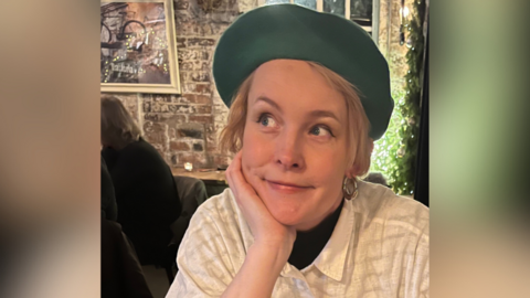 A photograph of Tracey Meek, a white woman with short blonde hair wearing a green beret and gold earrings and leaning on her hand while looking to her left. 