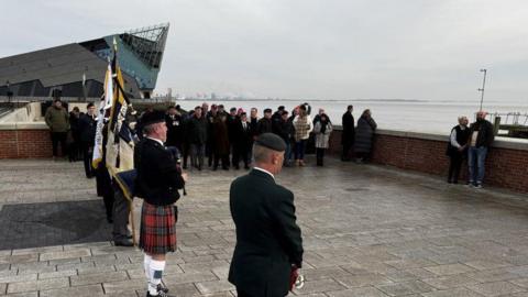 A group of people gathered at the ceremony on a paved area near The Deep in Hull. They are dressed smartly. A bagpiper can be seen in the foreground wearing a kilt. Several flagbearers stand next to him. The Humber estuary is in the background. 