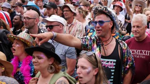 Man wearing a sparkly top in a crowd of people pointing to the left hand side of the photo