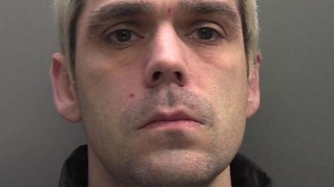 A police mugshot of a man with short, bleached blond hair looking into the camera. You can see the collar of his black padded coat.