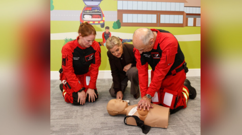 Two crew members of the Thames Valley Air Ambulance performing a CPR training on a manikin. Charity Royal Patron, The Duchess of Edinburgh, is watching them. They are kneeling in front of a colourful wall. The manikin is of a woman. The black bra across its chest has been cut.