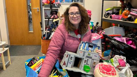 A woman with curly brown hair and glasses is smiling at the camera. She is wearing a pink puffer jacket and is holding children's toys in her hand while sitting in the floor