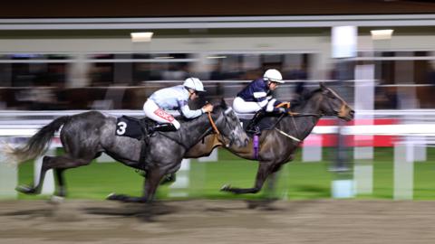A side-on view of two jockeys on two horses racing on a sandy track, with a crowd behind them which is out of focus.