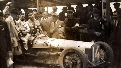 A sepia photo of men around a old racing car.
