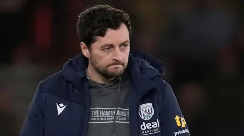 A man with dark hair and trimmed beard looks pensive at a football ground. He is wearing a dark tracksuit top